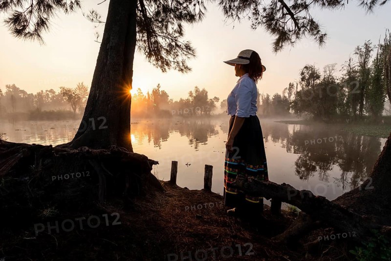 A woman in traditional attire stands by the water in Xochimilco, Mexico, as the sun rises, illuminating the misty landscape with a warm glow, showcasing cultural beauty