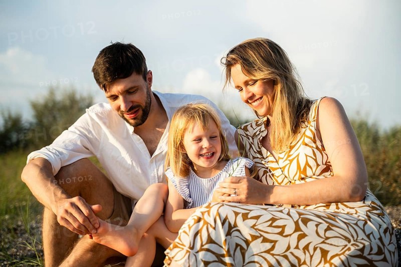 A joyful family sits together on an Italian beach, basking in the sun. The parents share a tender moment with their smiling daughter, surrounded by nature's beauty