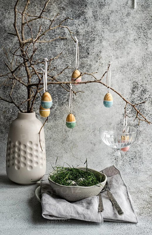 An Easter table setting featuring a ceramic vase with suspended painted eggs from bare tree branches and a bowl of grass accented by subtle textures of a tablecloth