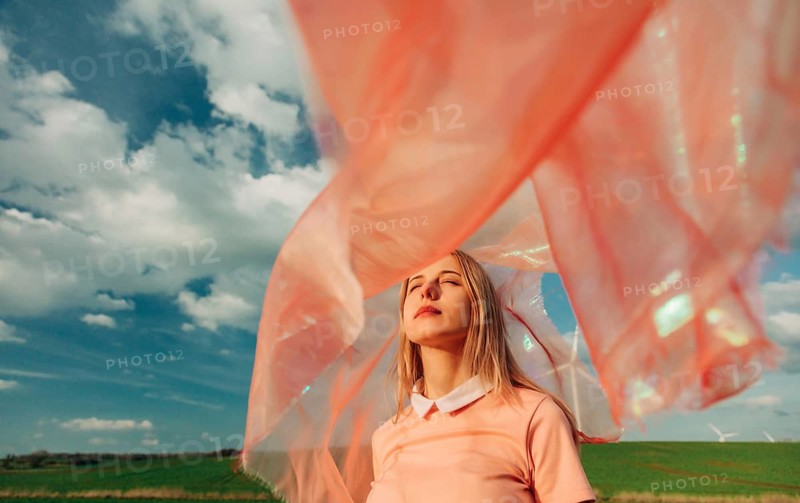 A woman stands in a field with her eyes closed, enveloped by a flowing pink fabric. The sky is bright with clouds, capturing a serene and tranquil moment