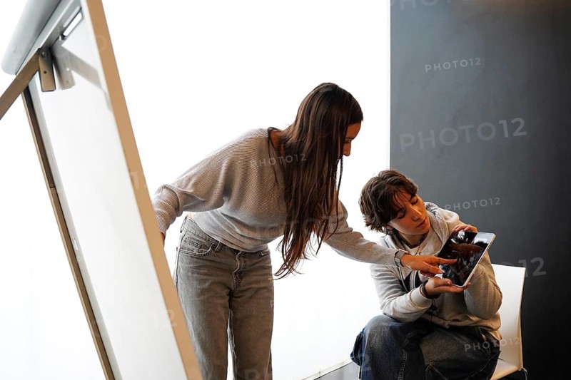 Two teenagers, a non-binary individual and a female, are fully engaged in an educational task, exploring digital content on a tablet in a school environment. They are collaboratively focused on the tablet screen, promoting an atmosphere of learning and companionship