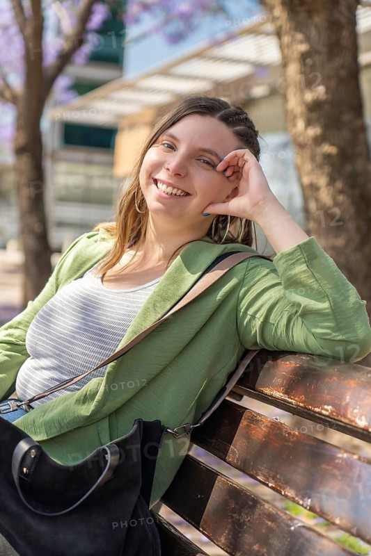 A cheerful woman in a green jacket smiles at the camera while sitting on a bench in a sunny park lined with blooming purple trees. Blurred people walk in the background, enjoying the day