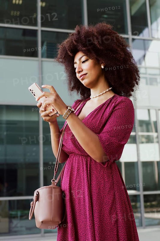A young Arab woman with afro hair and in a stylish pink dress uses her smartphone while standing outside a modern building, showcasing a blend of technology and fashion