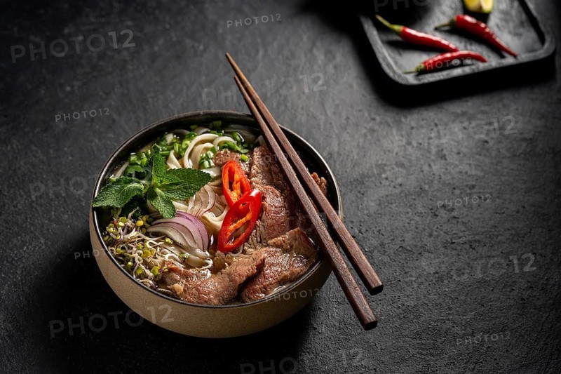 Taditional Vietnamese Beef Pho Noodle Soup served in a stylish dark bowl, garnished with slices of beef, red chili, onion, and fresh herbs on a dark background