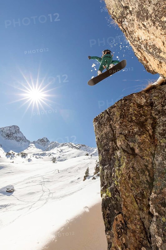 A snowboarder executes a thrilling jump off a rocky cliff, soaring through clear blue skies in a beautiful backcountry setting. Snowy mountains and sunshine enhance the dramatic scene