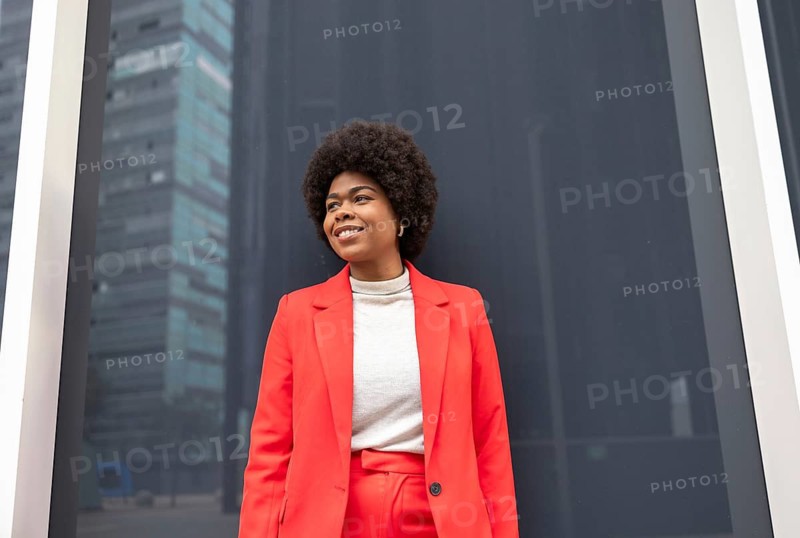 A woman in a bright red suit stands confidently against a modern urban backdrop. Her smile and posture convey empowerment and style, highlighting a vibrant lifestyle