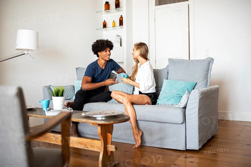 A young couple sits on a cozy sofa engaged in a cheerful conversation. The living room setting is modern and inviting, with pillows and decor enhancing the warm atmosphere