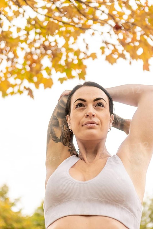 A woman stretches her arms while exercising outdoors during autumn in a New Zealand park. The vibrant fall foliage adds a scenic backdrop to her workout routine