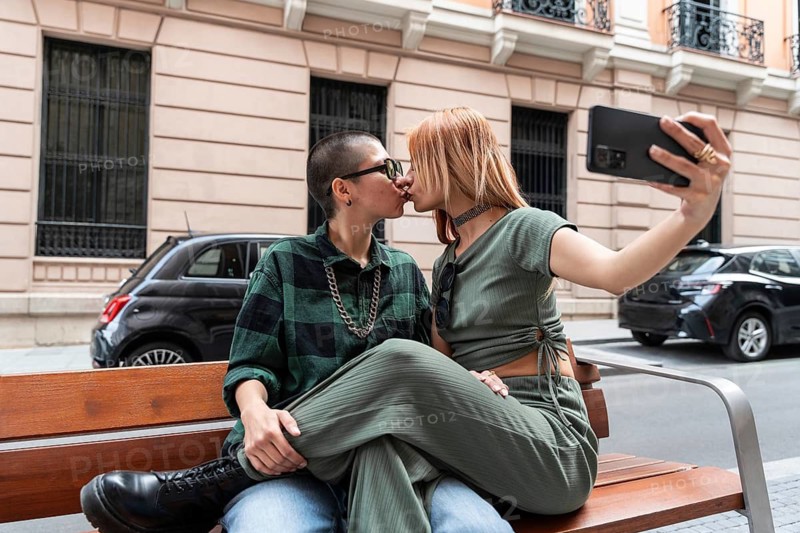 A Colombian lesbian couple captures a romantic moment with a selfie while kissing on a city bench. This image celebrates diversity, love, and the LGBTQIA+ community