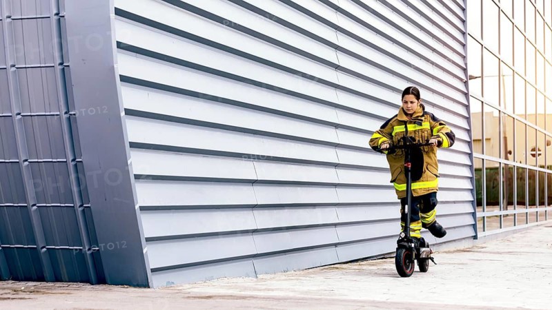 A girl wearing a firefighter suit rides an electric scooter against an urban backdrop with a modern architectural design, showcasing strength and confidence