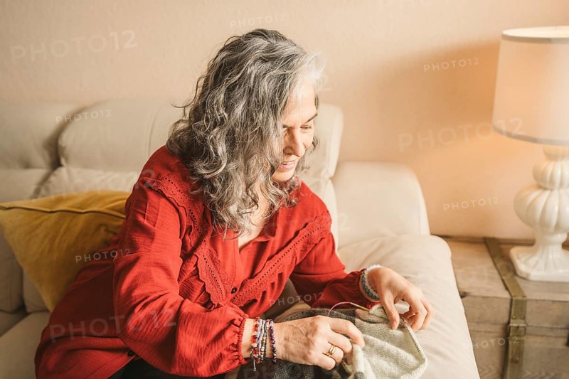 Elderly woman with gray hair sits on a beige couch, focused on sewing. A warm lamp light casts a soft ambiance in a cozy living room setting, enhancing the scene
