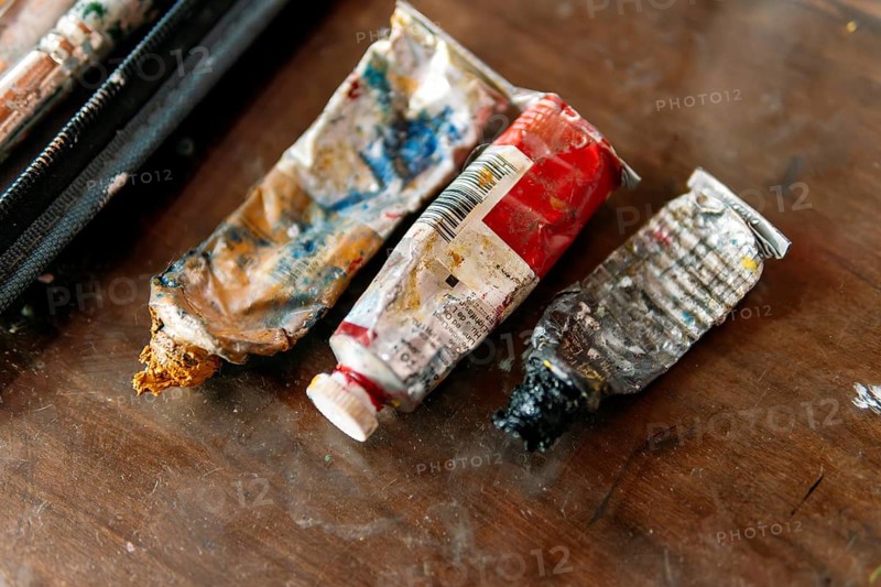 A close-up of worn and crumpled paint tubes covered in dried paint, resting on a wooden table. The messy textures and vibrant colors highlight the creative process of an artist