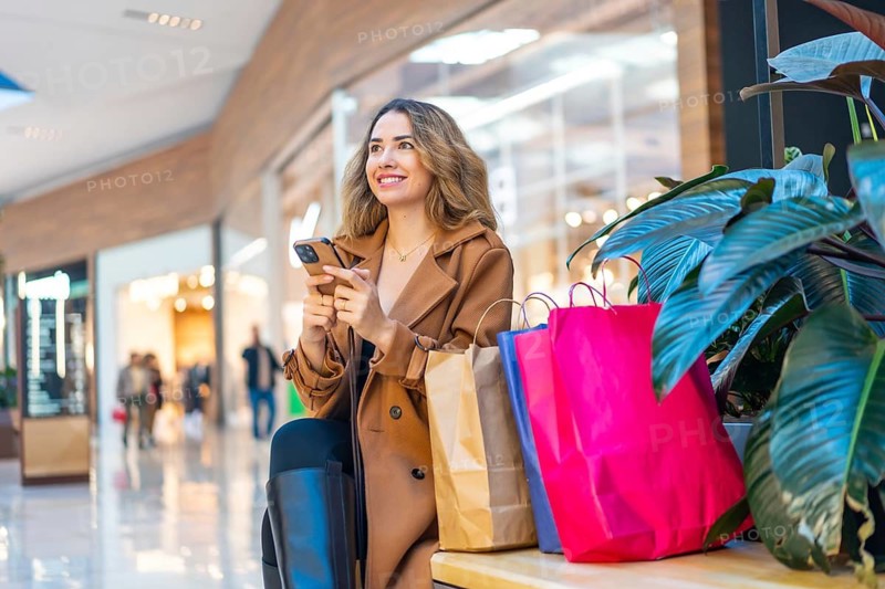 Happy woman holding a smartphone and shopping bags, enjoying a day of retail therapy in a modern shopping mall while looking around with a cheerful expression