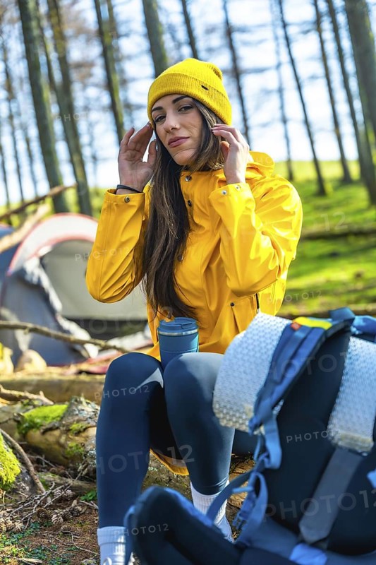 Young woman hiker adjusting her yellow beanie while sitting near her tent and backpack in a forest