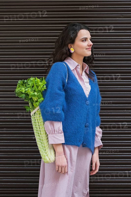 A stylish woman in a blue cardigan holds a net bag filled with fresh vegetables. She exudes confidence while showcasing sustainable shopping choices
