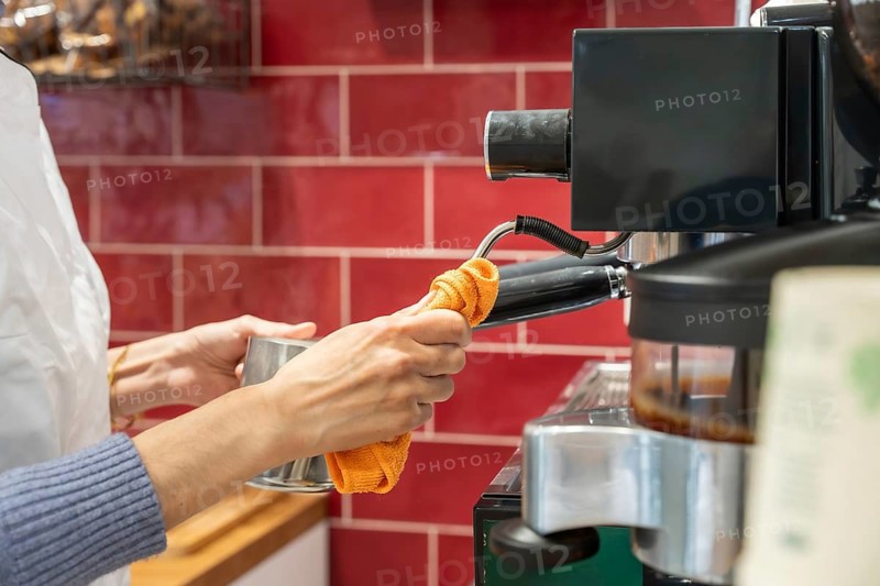 Barista wiping steam wand of espresso machine with orange cloth, preparing fresh milk froth, close up hygiene routine in a tidy cafe, professional service and attention to detail