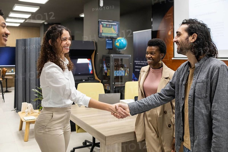 Colleagues greet each other warmly with a handshake in a modern office setting. The environment appears collaborative, with a cheerful exchange among diverse team members