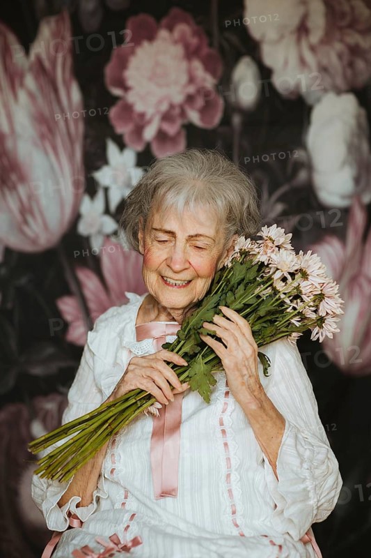 An elderly woman with a joyful expression holds a bouquet of flowers, surrounded by a floral backdrop. Her happiness radiates in this heartwarming studio portrait