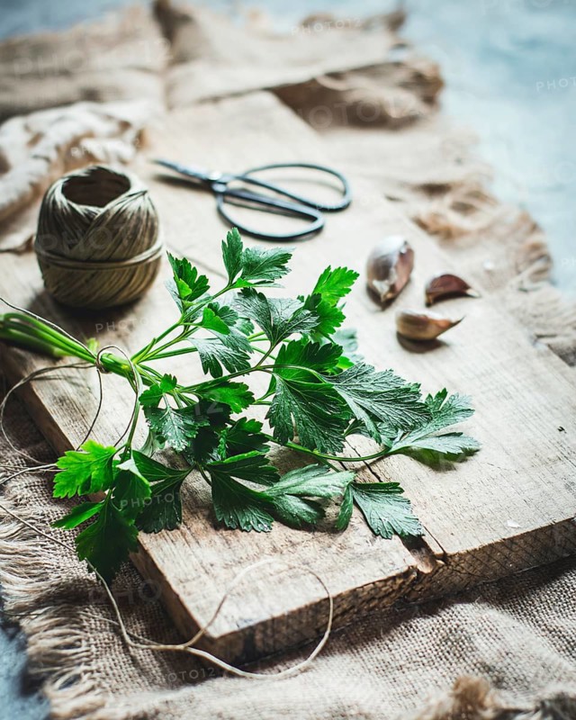 A bunch of fresh parsley lies on a rustic wooden cutting board, surrounded by garlic cloves, a spool of twine, and scissors, creating a cozy farm-to-table ambiance