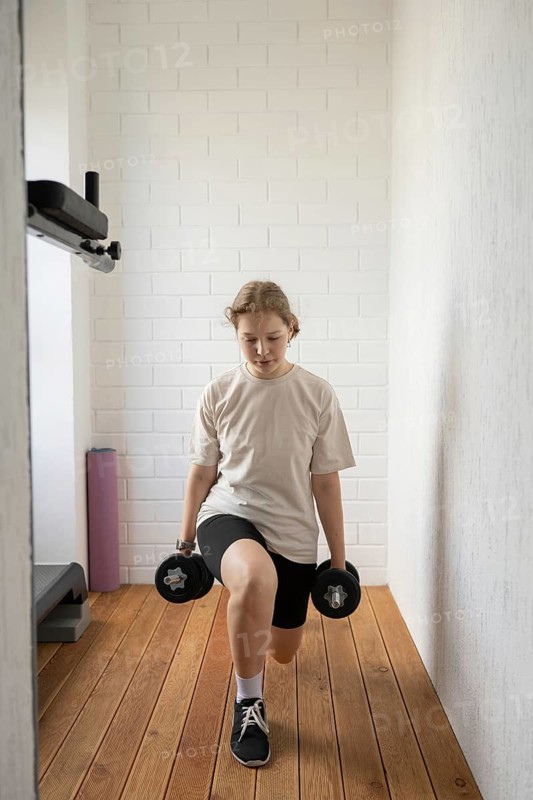 Teen girl practicing lunges with dumbbells in a bright home gym space. Wooden flooring and white brick walls create a cozy and motivating workout environment