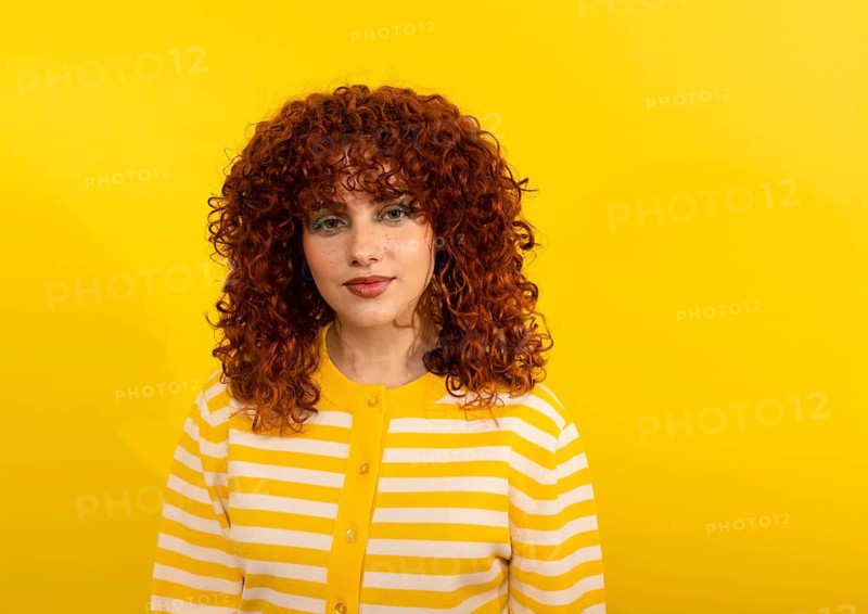 Portrait of a woman with vibrant, curly red hair wearing a yellow and white striped sweater, set against a vivid yellow background, casting harmonious and cheerful vibes