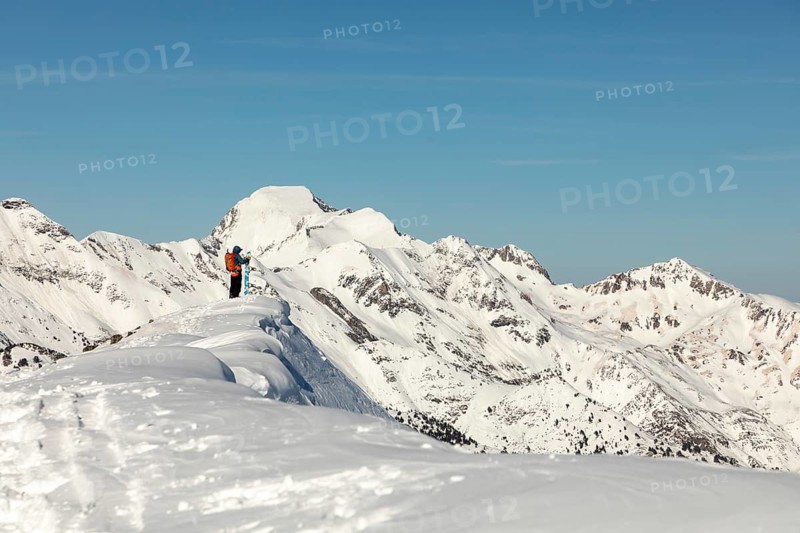A snowboarder with a bright orange backpack stands atop a snowy mountain ridge, overlooking a vast alpine landscape under a clear blue sky