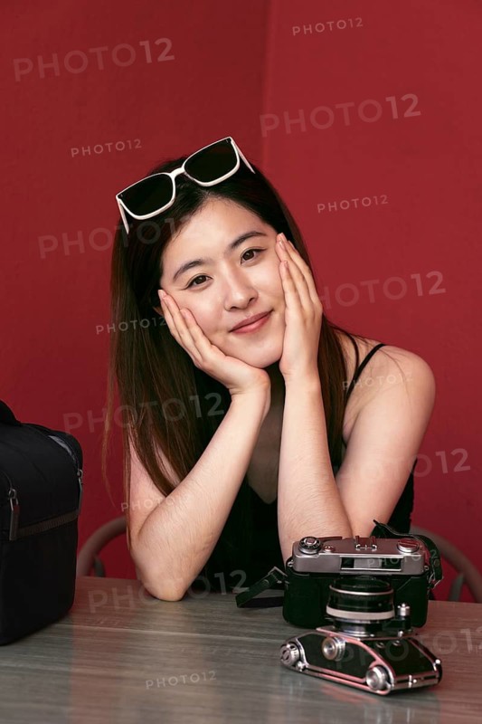 A young asian woman sits at a table with a vintage camera, sporting sunglasses on her head. The red background adds warmth to her cheerful expression, creating a friendly atmosphere
