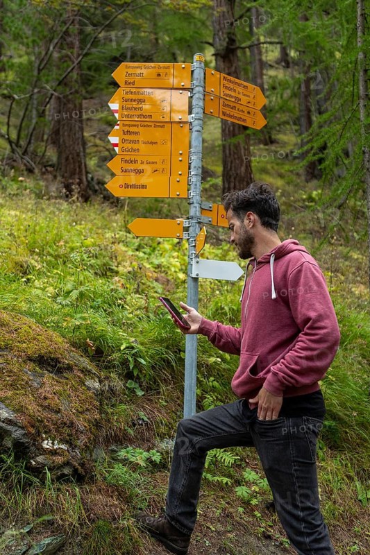 A man walks through a lush Swiss forest in autumn, consulting a smartphone. He wears a casual hoodie, surrounded by vibrant foliage and signposts, capturing the essence of exploration
