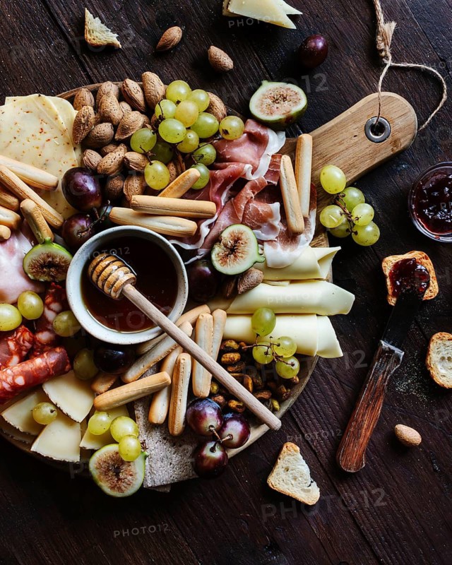 A rustic wooden board laden with an enticing variety of snacks including cheeses, grapes, figs, and breadsticks, complemented by a small bowl of honey, perfect for gatherings