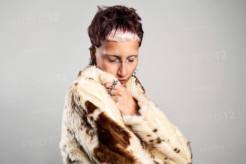 A woman with closed eyes in a unique hairstyle and white eyeliner wears a faux fur coat, accessorized with bold jewelry. The pose and expression convey confidence and individuality