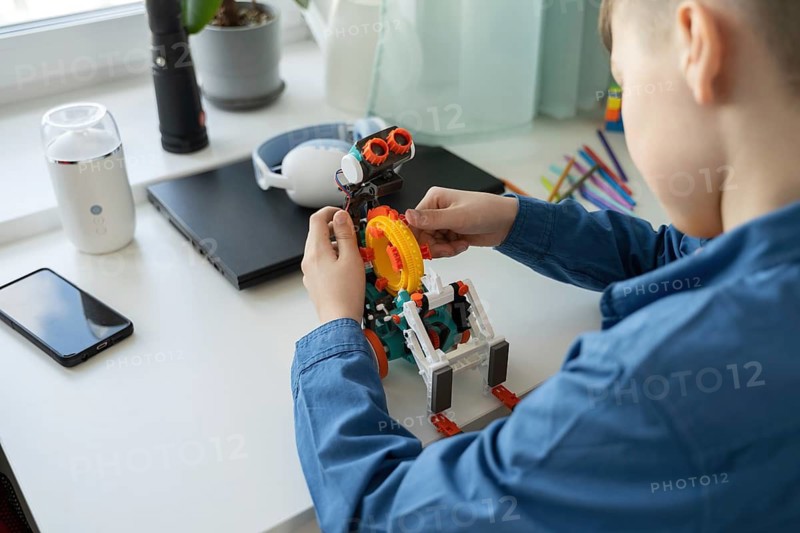 A young child focuses intently on assembling a colorful robotic toy on a desk. Surrounding him are various devices, fostering creativity and learning in a home environment