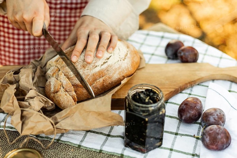 Hands slicing rustic bread on a wooden board, surrounded by fresh plums and jam. An inviting scene for preparing delicious homemade canapes in a cozy kitchen setting