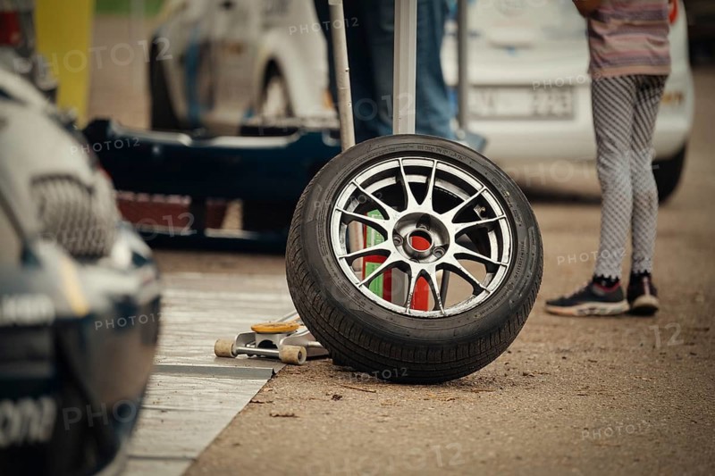A close-up of a racing tire resting on the ground in a motorsport pit stop area. The scene includes car parts, tools, and a blurred background of vehicles and people, capturing the dynamic atmosphere