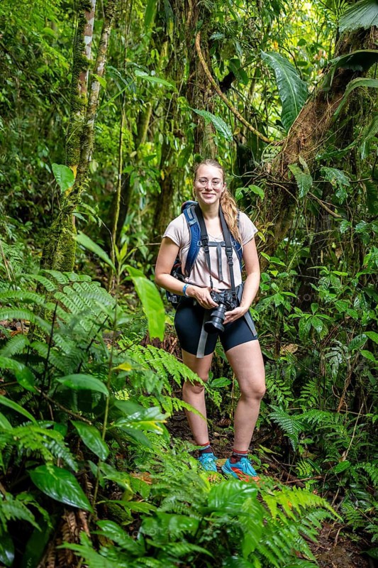 Young woman with camerea, tourist on a narrow hiking trail between dense vegetation in the tropical rainforest, Laguna de Hule, Refugio Nacional de Vida Silvestre Mixto Bosque Alegre, Alajuela province, Costa Rica
