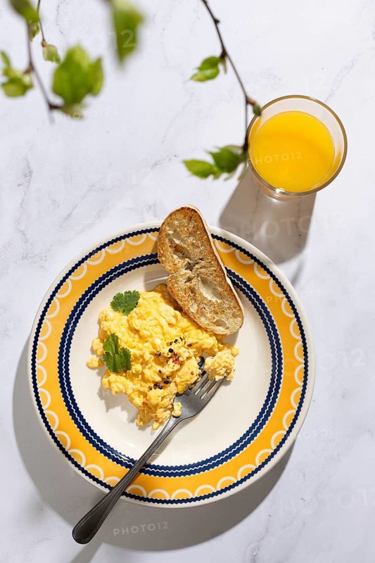 A delightful breakfast scene featuring scrambled eggs garnished with herbs, served alongside crispy toast on a vibrant plate. A glass of orange juice completes the meal, set on a white marble surface