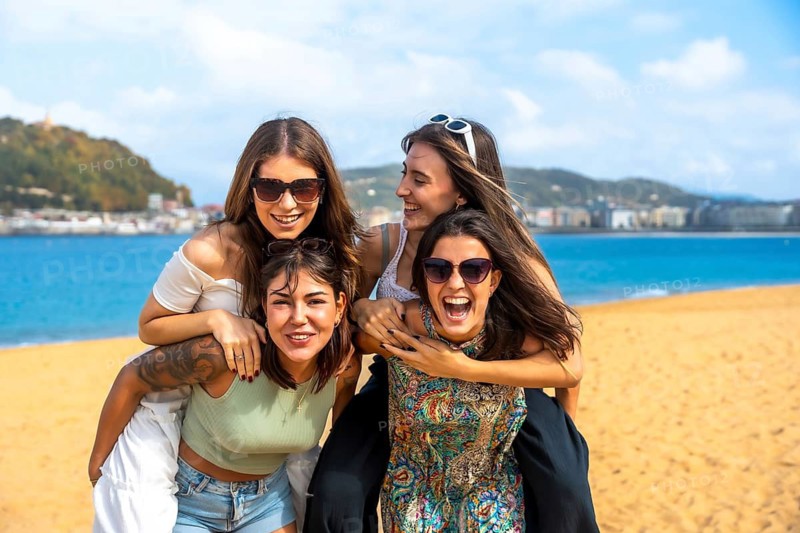 Four smiling young female friends laughing and carrying each other on a sandy beach, celebrating friendship and fun times during their summer holidays by the ocean