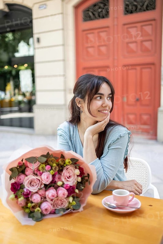 A woman sits at an outdoor cafe table, smiling with delight as she admires a beautiful bouquet of flowers purchased from a florist, enjoying a peaceful moment