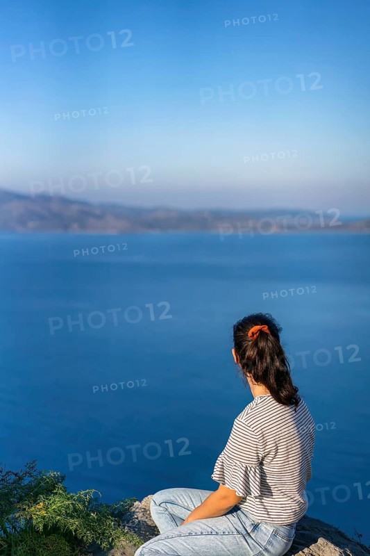A woman in casual attire sits on a rocky outcrop, gazing at the tranquil blue sea under a clear sky in Greece. The scene captures peaceful contemplation and natural beauty