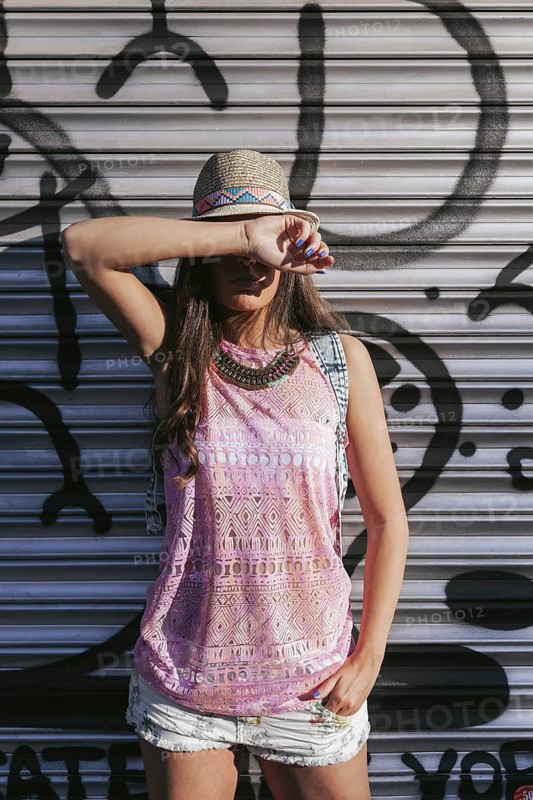 A fashionable woman poses against an urban backdrop, wearing a summer outfit with a hat and necklace. She covers part of her face, creating a mysterious vibe