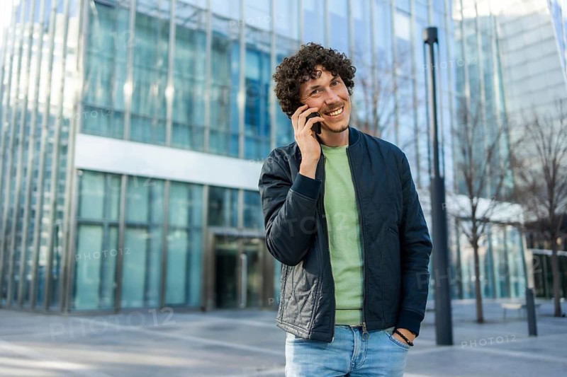 A businessman enjoys a phone call while standing outside a contemporary glass building. He wears a casual jacket and jeans, expressing happiness and connectivity