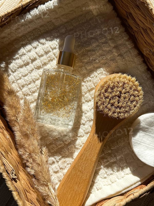 A beautifully arranged basket featuring a gold infused serum cosmetic bottle, a wooden brush, and soft towels, bathed in warm natural light in the bathroom. Ideal for wellness or skincare themes
