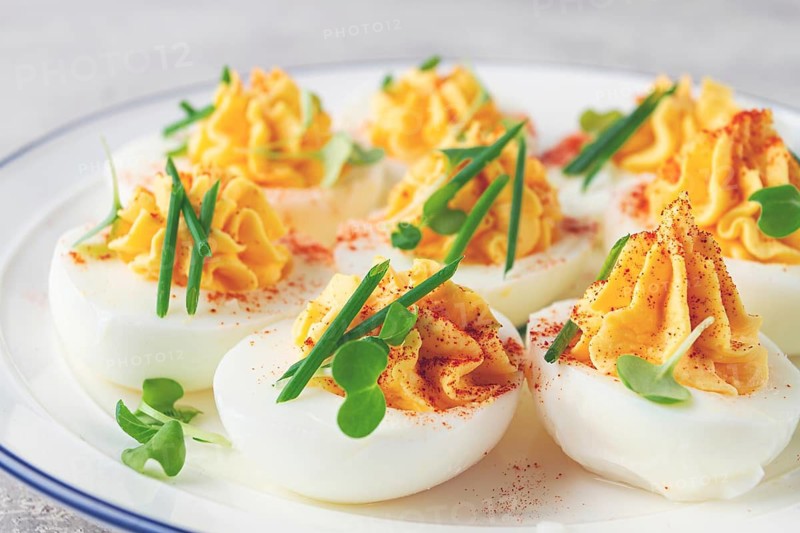 A plate holds several deviled eggs filled with yellow mixture and topped with spices and green garnishes. The dish is placed on a smooth light surface