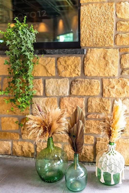 Elegant wedding decor featuring green glass vases, pampas grass, and ivy against a stone wall, creating a rustic and natural ambiance