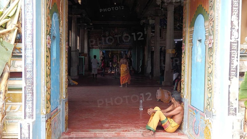 A man sits inside a colorful temple while others move in the background, Sri Lanka