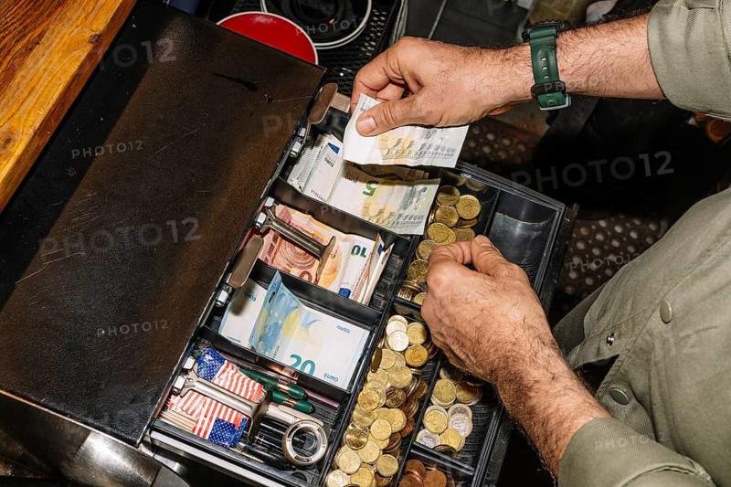 A barista or cashier handles euro banknotes and coins in an open cash register inside a coffee shop. The organized till contains currency, receipts, and small accessories on a wooden counter
