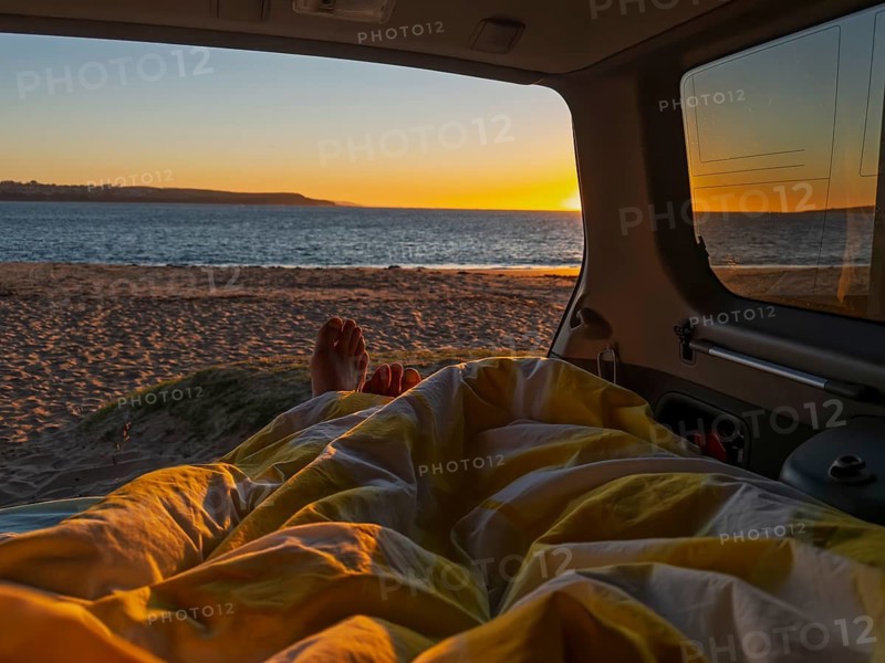 The photo captures a serene sunrise view from inside a camper van in Phillip Island, Australia. A cozy bed offers comfort as golden sunlight illuminates the sea, creating a tranquil beach escape