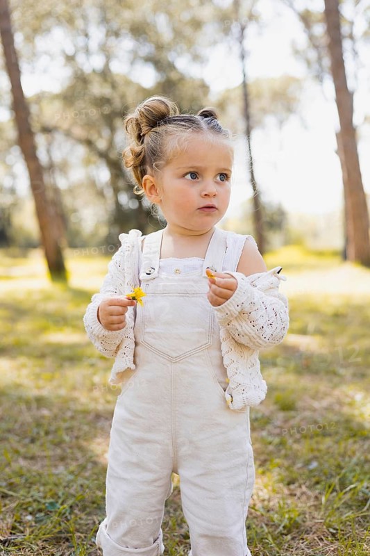 A young girl stands in a sunny forest, holding a small yellow flower. She wears a white knit sweater and overalls, enjoying the peaceful and serene outdoor environment