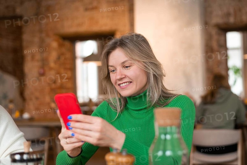 A woman in a green sweater is smiling while looking at her smartphone in a cozy cafe. She enjoys a peaceful breakfast moment surrounded by a warm atmosphere