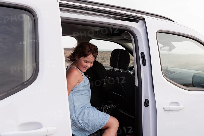 A joyful child in a light summer dress steps out of a parked car, ready for a fun-filled summer adventure. The open road awaits under a sunny sky, promising new experiences