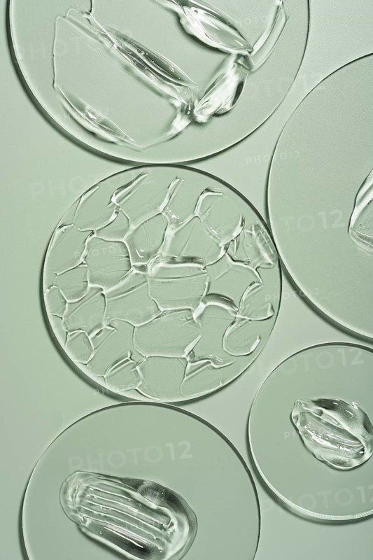 Top view of transparent gel smears on laboratory glass arranged over a soft green background, showcasing textures and fluidity used in skincare and cosmetic analysis